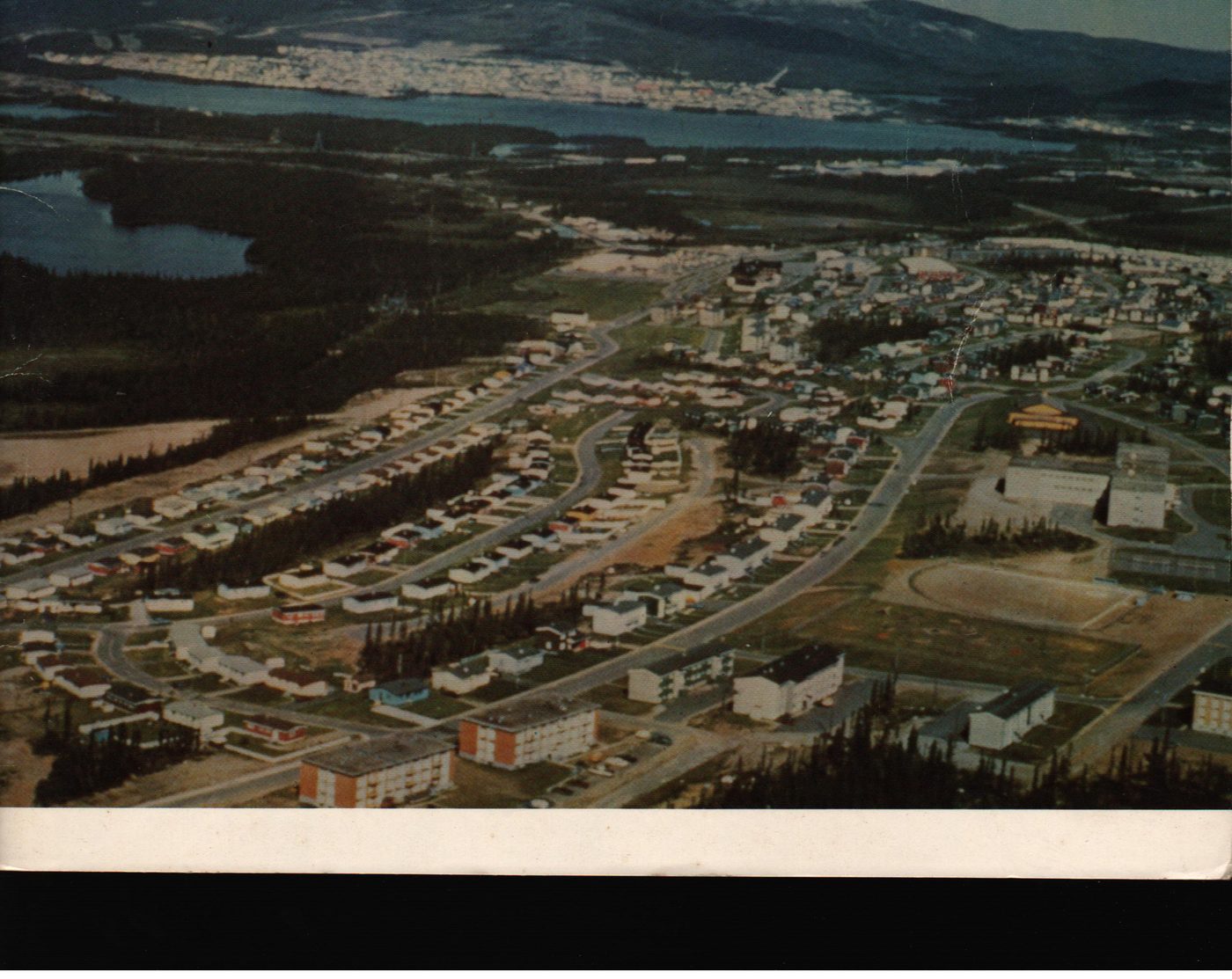Aerial colour photograph of Wabush in the foreground with rows of bungalows, curved streets, and community buildings, and Labrador City visible as a white sprawl across the far hillside